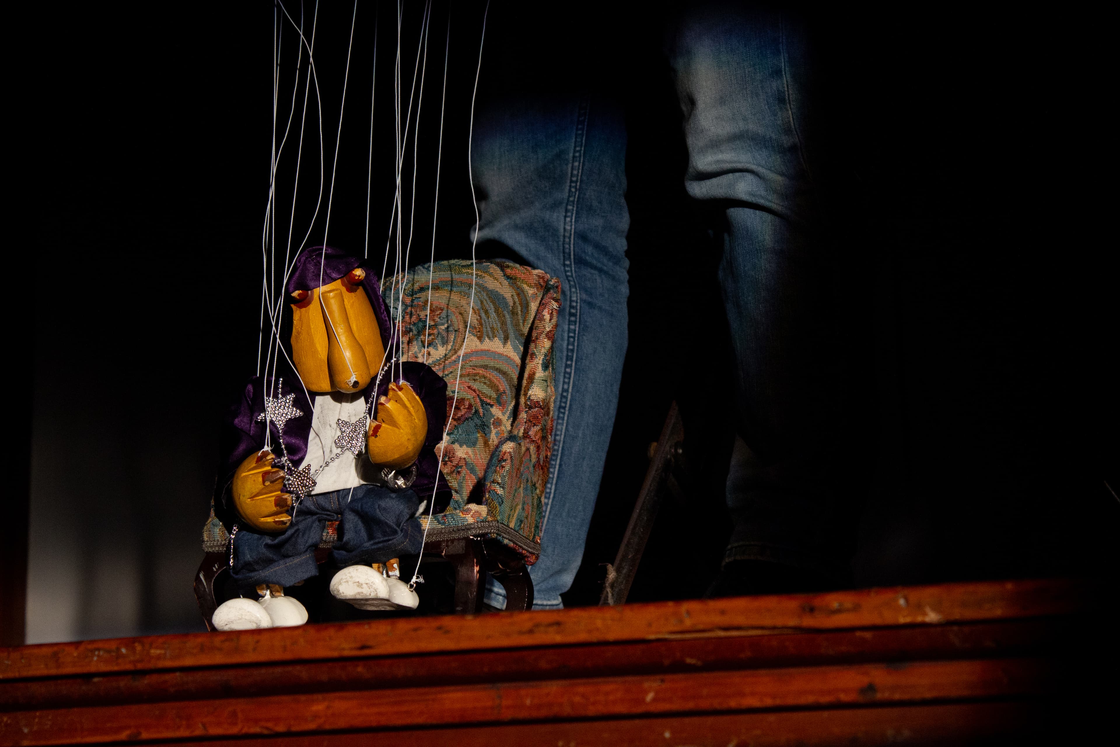 a wooden string puppet wearing a purple velvet hoodie, white tee-shirt silver star necklace, blue jeans and white shoes and sits in a chair. He has a stylised cylindrical face with a long nose and two red beady wooden eyes protruding just below the hoodie. The puppet's head is slightly tilted one hand is gesturing towards the audience. The strings and puppeteers legs are visible.